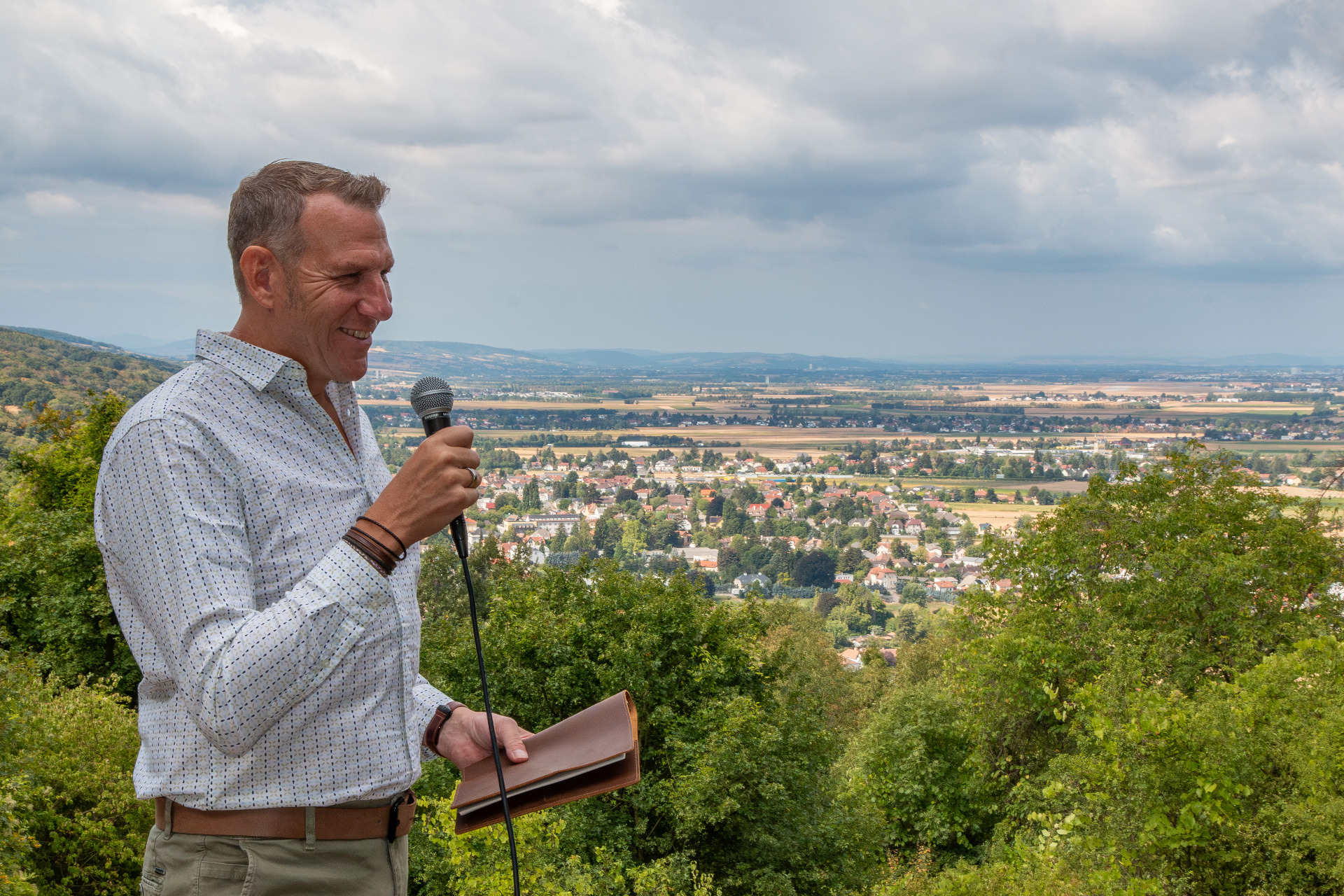 Mann mit Mikrofon hält Traurede, blickt lächelnd auf eine Stadtlandschaft unter bewölktem Himmel.
